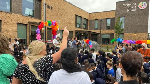 BBC School children dance to a brass band along with teachers, colourful balloons decorate the front of the new school