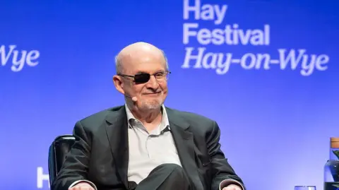 Getty Images Salman Rushdie at the Hay Festival, smiling at the audience, wearing a light grey shirt and a black suit.