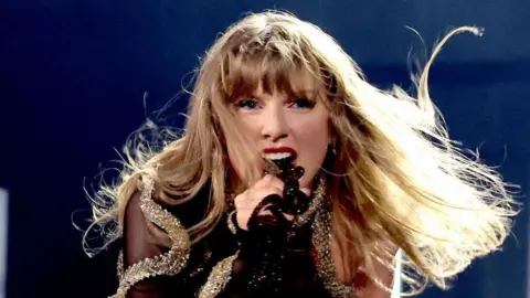 Getty Images Taylor Swift performs on stage in December last year in Vancouver, Canada, as part of her Eras tour. She is singing passionately into the microphone and is wearing a black and gold sequinned outfit.
