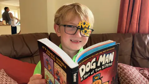 Vanessa Clarke / BBC Harry is sitting on a couch reading a book called Dog Man. He is smiling at the camera wearing glasses and a patch over one eye with rockets and stars on it.