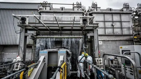 BBC The Magnox Swarf Storage Silo, a futuristic looking factory type building at Sellafield. Two people in yellow helmets and hazard protection suits are working with their backs to the camera. The building is metal and concrete with no windows or obvious openings. Pipe work and industrial equipment can be seen all over the image.