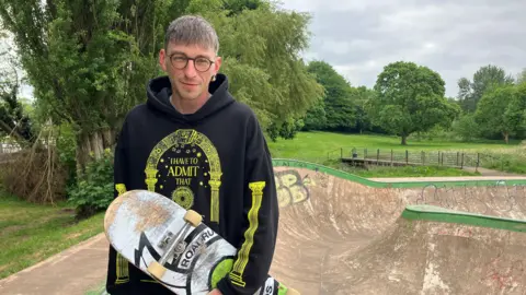 BBC A man wearing a black hoodie with yellow detailing is holding a skateboard and is stood at the top of a skateboard park. There are fields and trees in the distance.
