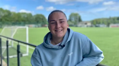 BBC Chloe is standing on the side lines of a football pitch wearing a blue hoodie, her hair is tied back and she is smiling at the camera. There is a football goal in the background.