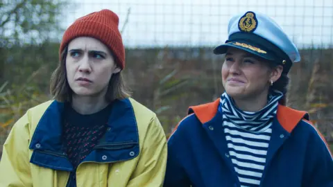 BBC/Various Artists Limited Two women sit next to each other outside. One is wearing a yellow rain mac with an orange beanie hat and the other is wearing a striped top with a blue jacket and sea captain's hat.