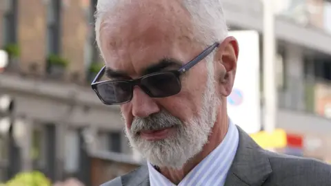 PA Media A close up of the face of Cdr Julian Bennett, wearing sunglasses - he has a grey beard and grey hair and he is looking down.