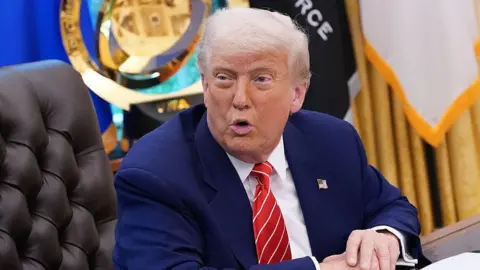 Getty Images US President Donald Trump sits at a desk in the Oval Office of the White House.  He is wearing a blue suit with an American flag pinned to the lapel. 