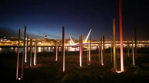 Getty Images a bridge is illuminated behind a light sculpture made up of steel columns entitled on the banks of a river. a number of buildings can be seen in the background