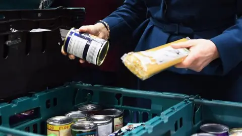 PA Media A woman holds a packet of spaghetti in her left hand and a tin in the right, above a stack of plastic food baskets.
