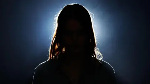 Getty Images An anonymous female figure. We can make out shoulders and long hair. There is a bright white light behind her on a black background. 