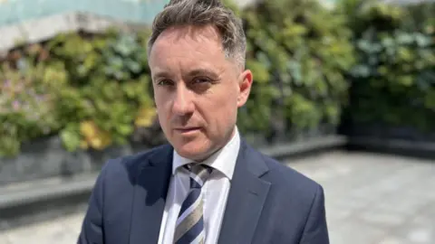 Tom Fletcher in a smart blue suit. He has a blue and gold tie on. He has short hair and is clean shaven. The background is blurred but we can see some bushes behind him. 