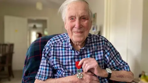 Alex Pope/BBC Leslie Lemon, holding a Légion d'honneur medal in his hands, wearing a blue checked shirt, sitting in a chair, with short grey hair. He has a watch on his left wrist. 