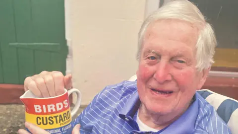 Supplied Leslie aged 100, holding a Bird's Custard jug, in a chair, with short grey hair, and wearing a blue and white top. He is smiling, and a green door is behind him. 