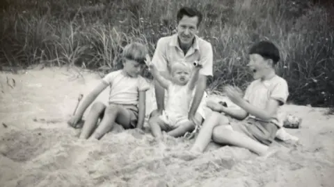 Supplied Leslie on a beach with his three children, Richard, Mary and Michael. Leslie is looking at the camera, and Mary has her arms up, with Michael looking at her and clapping. The image is black and white. 