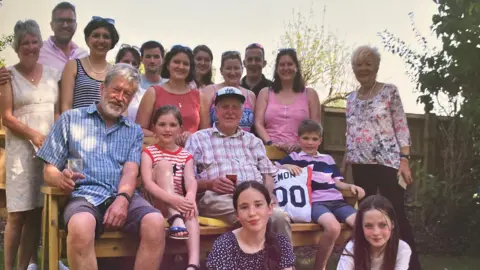 Supplied An image of a large family photo, including several children, some sitting on the floor, others on a bench. They are all smiling and looking at the camera. They are outside, by a fence with a tree to the right. 