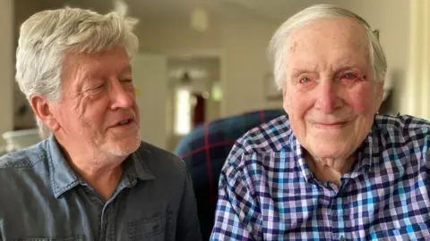 Alex Pope/BBC Richard, looking at his father, Leslie. He is wearing a blue plain shirt. His father is wearing a blue checked shirt. They both have short grey hair and are smiling. 