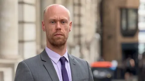 A man in a grey suit and purple shirt and tie stands outside a white stone building. 