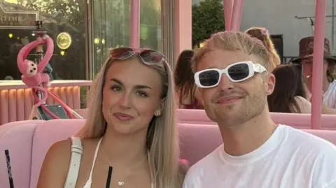 Family photo Kai Roberts with his girlfriend Kya Gillane-Heywood sitting on a pink sofa and smiling at the camera.