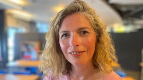 Dr Lindsey Cameron looks into the camera - she's sat in a school room and is wearing a pink T-shirt. She has blonde curly hair.