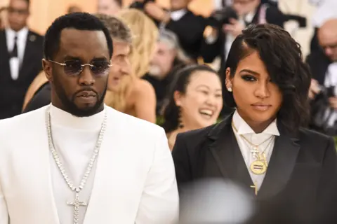 Getty Images Sean Combs and Casandra Ventura, dressed in formal attire: Combs in a white suit with an ornate cross necklace, Ventura in a black suit with layered necklaces including a circular pendant and a cross.