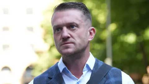 PA Media Tommy Robinson is wearing a light blue shirt and a checked blue waistcoat with a messenger bag over his shoulder. He has short hair and light stubble.