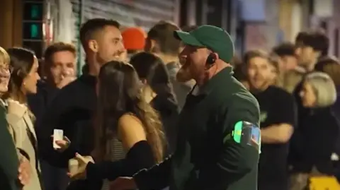 GMP Image shows people queuing for a club in Manchester and a member of a door staff team talking to some of the people in the line 