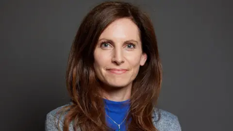 UK Parliament Chester South and Eddisbury MP Aphra Brandreth. She has long auburn hair and is wearing a blue sweater under a grey jacket. 