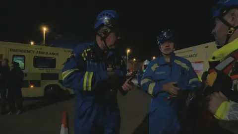 HM Coastguard Fleetwood HM Coastguard rescuers discuss the plan during a major search. Two men in blue suits with yellow reflective patches stand in front of ambulances. 