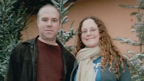 Rob Webb A man with brown hair wearing a maroon jumper and black denim jacket stands next to a woman with brown long curly hair wearing a blue shirt and cream scarf.  They are smiling and stood in front of some Christmas trees with snow on the branches.