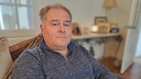 A man with short grey hair is looking at the camera.  He is wearing a blue zip jumper and behind him is a table with photos.