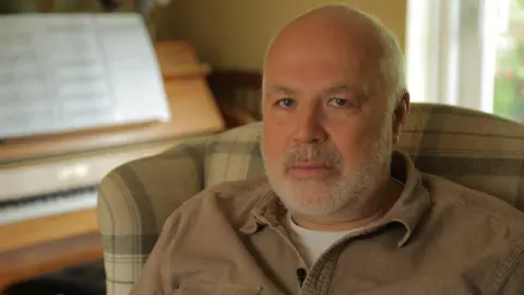 A man with shaved head and short beard loosk straight at the camer.  He is wearing a brown shirt and is in an armchair.  A piano can be seen in the background.