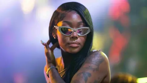 Getty Images Summer Walker wears a one-armed black top, revealing a heavily tattooed left arm. She has bobbed black hair and wears reflective sunglasses, her left hand running through her hair. 