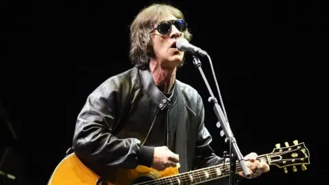 PA Media Richard Ashcroft on stage at the Principality Stadium, Cardiff, one of the support acts for Oasis as the band's long-awaited reunion tour kicks off in Wales. He is holding a guitar and leaning towards a microphone. He is wearing sunglasses and a leather jacket. 