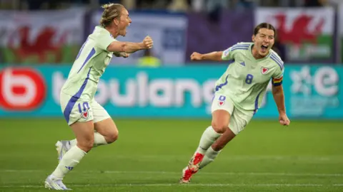 FAW Jess Fishlock with clenched fists cheers and runs in celebration after scoring, while teammate Angharad James with arms out runs ahead of her. Wales flags out of cous in the background. They wear Wales' lime green away kits.