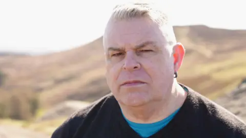 BBC Wales Alisdair does not smile as he stands near some rocks with a hill behind him. He is wearing a bright blue t-shirt under a dark v-neck jumper and has an earing in his left ear. His short white hair is brushed back.