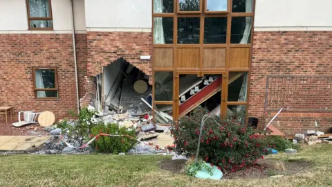 A large hole at the bottom left of a brick and white stone building next to a window. Bricks and debris from the wall are scattered all around and in the bushes and grass in front of the building. 