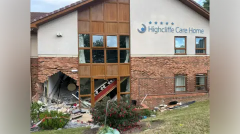 A brick and stone building with the sign reading 'Highcliffe Care Home' in blue writing. A large hole can be seen at the bottom left of the building next to a window. Bricks and debris from the wall are scattered all around and in the bushes and grass in front of the building. 