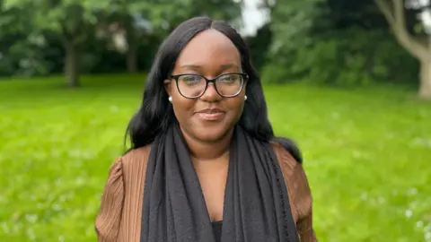 Aaliyah is in a park,  standing on the grass in front of some trees and opposite a playground area