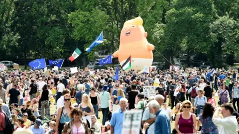 PA Media A large inflatable doll of a baby with a blonde quiff and wearing a nappy is held above a crowd of people in a park. There are several flags being flown, including a saltire and European Union flags. 