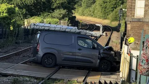Lauren Burnham A dark grey van which has crashed into a level crossing. The bonnet of the van is smashed.