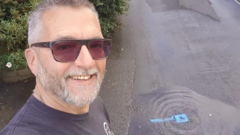 Dean Majors A man with grey hair and a beard, wearing sunglasses, takes a selfie next to a flooded area of road with the leak point marked in blue. 