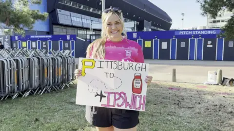 Cameron Noble/BBC Morgan Summersgill standing outside Portman Road football stadium. She is holding a sign that says Pittsburgh to Ipswich on it. She is wearing a pink Ipswich Town football shirt and smiling while looking into the camera. 