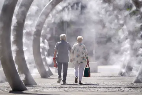 PA Media A man and a woman - both with silver hair - are walking inside a row of ring metal structures that are spraying mist on a very sunny day. A man is wearing a checkered blue shirt with short sleeves, jeans and trainers and a woman in white trousers and a loose-fitting shirt.
