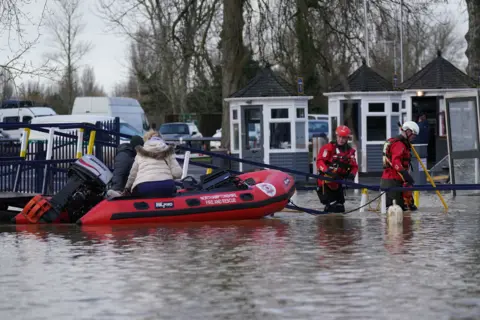 PA Media Two men from a fire and rescue service wearing red gear and helmets are wading in the flooded water and dragging a boat with two people on it. The water comes up to just above the knee level of the two men. We can not see the faces of the two people on the boat but one has a white winter coat and has blond hair tied at the back.