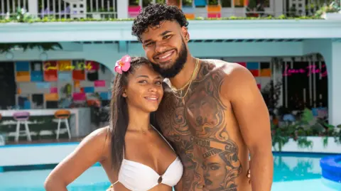 Kim Nunneley/Peacock via Getty Images Amaya Espinal and Bryan Arenales pictured in the Love Island USA villa. Amaya has her brown hair pinned back with a pink flower and poses with her hand on her hip while wearing a white bikini. Bryan leans into Amaya smiling. He has curly dark hair and a short beard. He is topless, revealing a heavily tattooed chest with images of wolves, women and caution tape. They're pictured in front of a swimming pool and bar. 