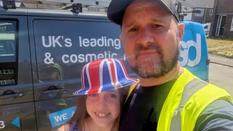 Stuart Field Stuart Field and his daughter take a selfie next to a grey van. He is wearing a high-vis jacket and a black cap. His daughter is seen hugging him while wearing a union jack bowler hat.
