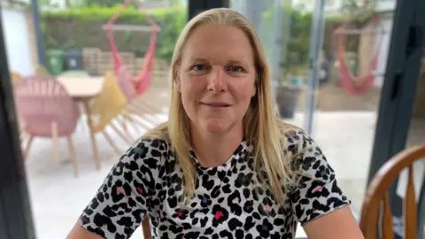 A woman in her early 50s with shoulder length blond hair smiling and wearing a colourful animal print t-shirt sat at a kitchen table. 