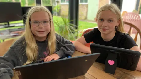 Rosa is an 8 year old girl with long blond hair and wearing glasses. She is wearing a grey jumper and smiling. On her righthand side is 14 year old sister Matilda with long blond hair, wearing silver ear rings and a black T-shirt. Both are sat at a table with devices in front of them and smiling. 