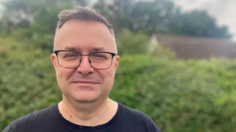 A man looking at the camera. He is wearing glasses and black T-shirt. A hedge is behind him.