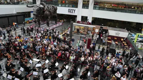 PA Media A large orchestra performing in front of a large crowd next to a large bull statue at a train station.