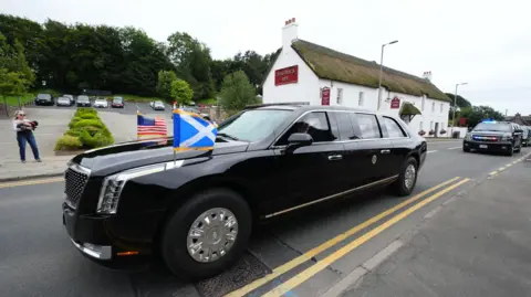PA Media The presidential car with the flags of the US and Scotland flying from the bonnet passed through a Scottish village - a low building with a thatched roof is in the background. 